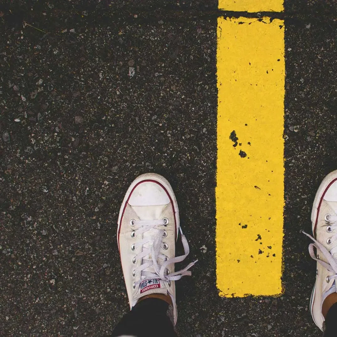 Artikelbild Startseite Deine Herausforderung Weiße Sneakers stehen vor einer gelben Straßenmarkierung auf asphaltierter Fläche.