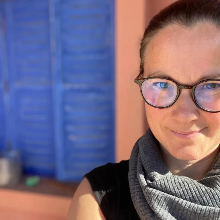 Artikelbild ueber mich Vanessa Lagnach mit Brille und Schal steht vor einer blauen Fensterläden-Wand.