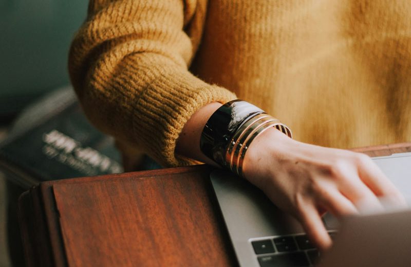 Frau mit gelbem Pulli arbeitet am Laptop Eine Frau tippt auf einem Laptop, trägt einen gelben Pulli und mehrere Armbänder.