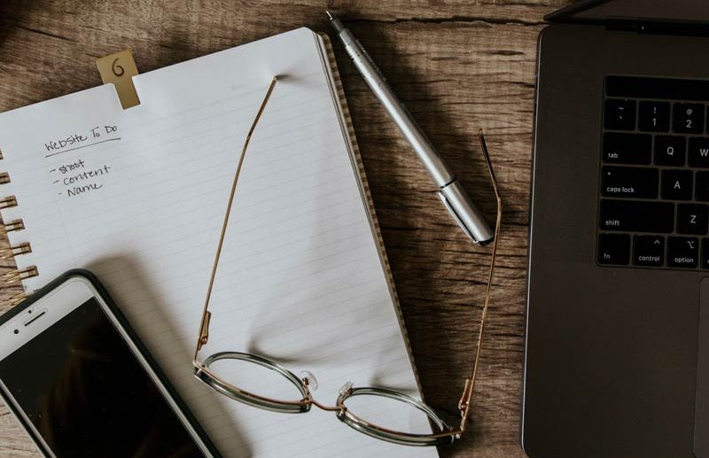 Arbeitsmaterial liegt auf einem Tisch Schreibtisch mit Notizbuch, Smartphone, Brille, Stift und Laptop, um damit eine Website zu planen.